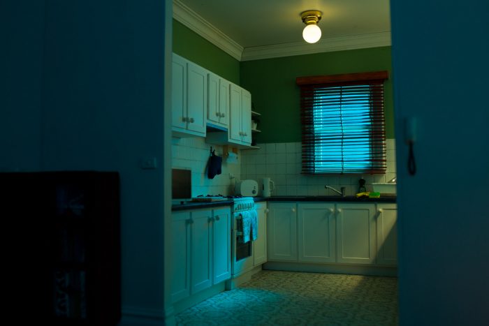 A miniature suburban kitchen at night, lit with green and blue tones. Eerie, empty.