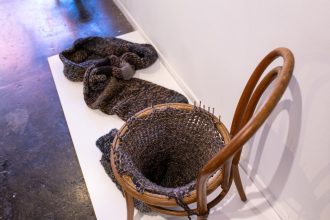 A sculpture where a Bentwood chair has been turned into a circular loom, with a dark knitted tube emerging from the bottom.