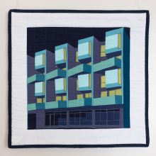 A figurative quilt showing a geometric apartment block in blues and greens.