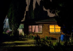 A suburban house photographed at night with dark shadows and two different coloured windows.